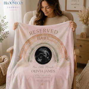 Puede incluir: Una suave manta rosa para bebé con un diseño de arcoíris y las palabras "RESERVED for our BABY". La manta presenta una imagen de ultrasonido y el nombre "OLIVIA JAMES". También se incluye el texto "After every storm comes a rainbow", junto con la fecha 05/05/2026.