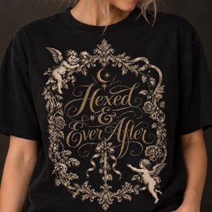 May include: A black short-sleeved t-shirt featuring a gold-toned graphic print. The design includes an ornate floral frame with two cherubs, a crescent moon, and a central dagger. The text "Hexed & Ever After" is written in elegant script.