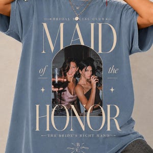Puede incluir: Una camiseta color azul jean con un estampado gráfico que incluye el texto "BRIDAL SOCIAL CLUB", "MAID of the HONOR" y "THE BRIDE'S RIGHT HAND - EST. 2026". El gráfico central muestra una fotografía de dos personas sosteniendo copas de vino.
