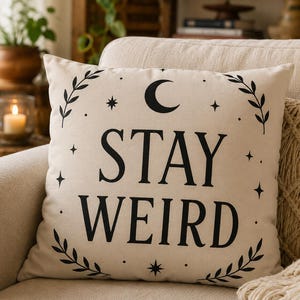 Puede incluir: Un cojín cuadrado de color blanco roto con el texto negro impreso STAY WEIRD en el centro. El diseño incluye una luna creciente negra, varias estrellas pequeñas y motivos de ramas con hojas que enmarcan el texto sobre la tela.