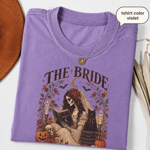 Puede incluir: Una camiseta violeta con un estampado gráfico de una novia esqueleto leyendo un libro titulado SPELLS, rodeada de flores, un gato negro, calabazas y velas. El texto THE BRIDE aparece impreso en una fuente oscura arqueada arriba.
