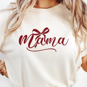 Könnte beinhalten: Cremefarbenes T-Shirt mit dem Wort "Mama" in bordeauxfarbener Schrift, verziert mit einer bordeauxfarbenen Schleife. Das Shirt hat kurze Ärmel und einen Rundhalsausschnitt.