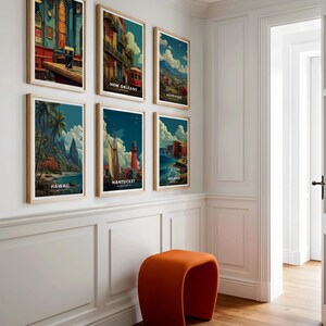 May include: A collection of six framed travel art prints featuring vintage-style illustrations of various locations, including New Orleans, Asheville, Hawaii, Nantucket, and Ireland. The prints are displayed on a white wall with an orange stool in front.