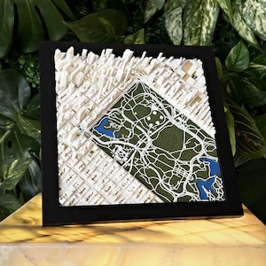 May include: A framed 3D topographic map featuring a white architectural model of city buildings alongside a green textured park area with blue water elements. The square artwork is encased in a simple black frame, displaying a detailed urban layout.