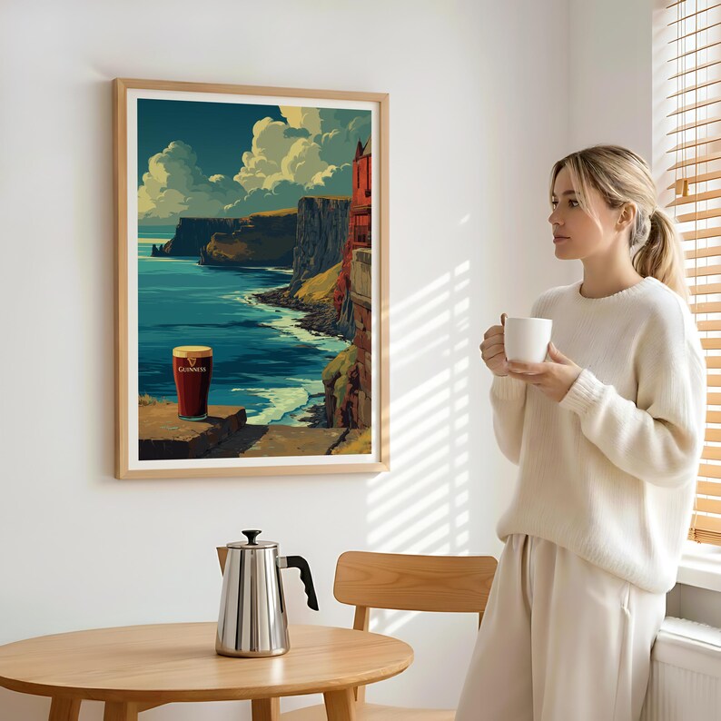 May include: A framed print of a coastal landscape with a pint of Guinness. The artwork depicts cliffs, ocean, and sky in shades of blue, green, and brown. A woman stands nearby, holding a white mug. A stainless steel kettle sits on a wooden table.
