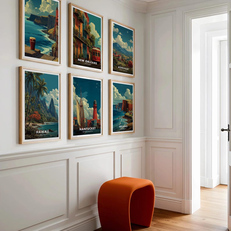 May include: Six framed travel posters with wooden frames are displayed on a white wall. The posters feature scenic views of Hawaii, Nantucket, Ireland, New Orleans, and Asheville. An orange stool sits below the posters. The room has white paneling and a doorway.