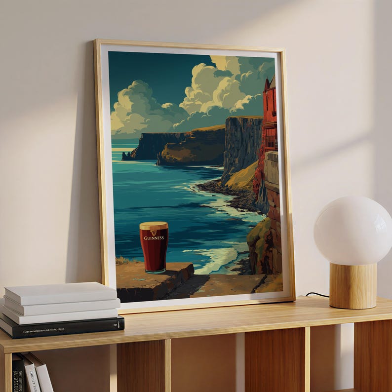 May include: Framed art print featuring a scenic coastal landscape with a pint of Guinness. The artwork depicts a dramatic cliffside view with a deep blue ocean, fluffy clouds, and a red building. The Guinness glass has a golden head.