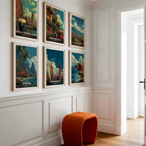 May include: Six framed travel art prints with wooden frames hang on a white wall. The prints feature vintage-style illustrations of various locations, including Hawaii, New Orleans, and Nantucket. An orange stool sits below the artwork.