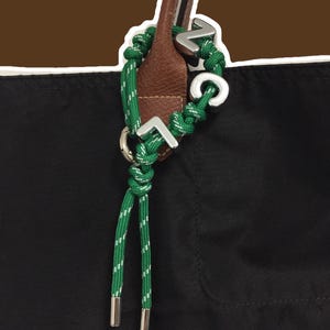 May include: A black tote bag with brown leather handles features a green rope charm with white reflective threading. The charm is adorned with silver-toned metal letters spelling out VCL, attached to a metal ring and finished with silver metal aglets.