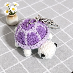 May include: Crocheted turtle keychain with a purple and white shell, white head and legs, and black eyes. The keychain has a silver clasp and key ring. A small vase with daisies is in the background.