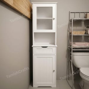 May include: A tall white bathroom cabinet featuring a top section with a glass-paneled door, an open middle shelf, a single drawer with a recessed handle, and a bottom section with a vertical-paneled door, each equipped with a small silver knob.