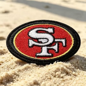 May include: An oval-shaped embroidered patch featuring a red background with a white interlocking SF logo in the centre. The design is bordered by a thin yellow line and a thick black outer edge, creating a distinct sports team emblem.
