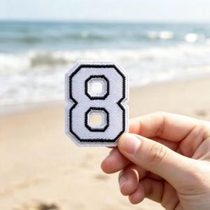May include: A white embroidered patch in the shape of the number eight, featuring a thick black border and a black inner outline. The patch has a textured fabric surface with visible stitching throughout the white and black sections.