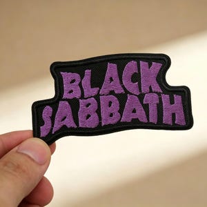 May include: Embroidered patch featuring the text "BLACK SABBATH" in purple, stitched onto a black background. The patch has a unique, curved shape and is likely designed to be ironed or sewn onto clothing or accessories.