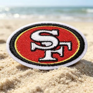 Puede incluir: Un parche bordado de forma ovalada que presenta el logotipo SF en blanco centrado sobre un fondo rojo. El diseño está rodeado por un borde amarillo fino, seguido de un borde negro grueso y rematado con un borde exterior blanco.