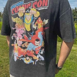 Może przedstawiać: Ciemnoszara koszulka z dzianiny wafelkowej z kolorową ilustracją komiksową i napisem "NEW YORK COMIC CON". Grafika przedstawia różnych superbohaterów w dynamicznych pozach.