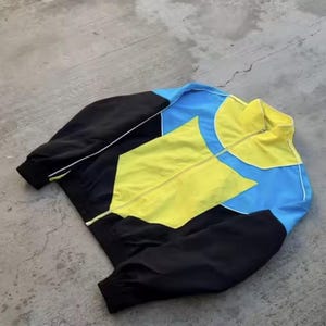 May include: A colour-blocked jacket in black, yellow, and blue. The jacket has a full zip, a stand-up collar, and white piping details. The sleeves and lower body are black, with yellow and blue geometric shapes on the chest and shoulders.