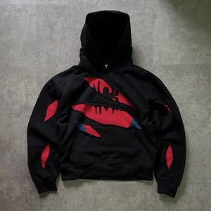 May include: Black hooded sweatshirt with a unique design featuring red and blue accents. The design appears to be torn or ripped, revealing a red graphic underneath. The sweatshirt has a front pocket and a drawstring hood.