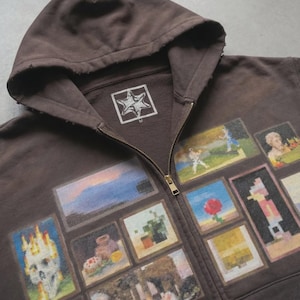 May include: A brown zip-up hoodie with a distressed appearance. It features a series of framed images on the front, including a skull, landscapes, and pixelated figures. The hoodie has a hood and a white logo on the chest.