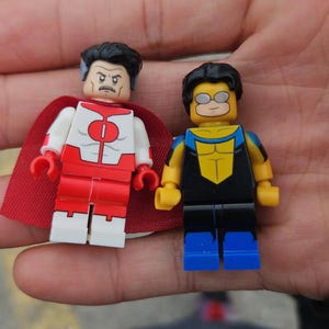 Puede incluir: Dos minifiguras de Lego de personajes de cómics. Una figura tiene un traje rojo y blanco con una capa roja. La otra figura tiene un traje negro y amarillo con detalles azules. Ambas figuras tienen rasgos faciales detallados.