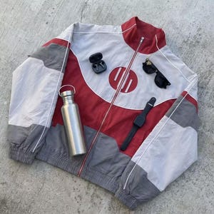 May include: A red, white, and gray track jacket with a full zipper, a silver water bottle, black sunglasses, a black smartwatch, and wireless earbuds. The jacket has a circular design on the front.