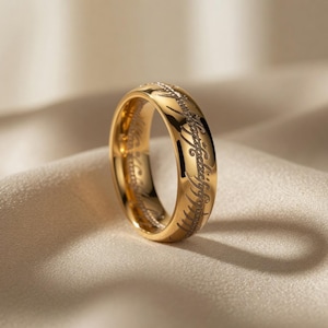 May include: A gold ring with engraved lettering. The ring is displayed on a neutral-coloured fabric. The text is in a stylized script, encircling the band. The ring's surface reflects light, emphasizing its metallic shine.