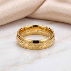 May include: A polished gold-toned band ring featuring an engraved script pattern that wraps around the entire outer surface. The metal has a smooth, reflective finish, and the intricate, flowing characters are etched into the gold-colored metal.
