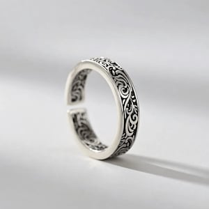 Könnte beinhalten: Ein verstellbarer Ring in Silber mit einem aufwendigen, wirbelnden Muster. Das offene Design ermöglicht eine Größenanpassung. Die Oberfläche des Rings ist mit einem dunklen, kontrastierenden Design versehen, das einen auffälligen visuellen Effekt erzeugt. Der Ring ist vor einem schlichten, hellen Hintergrund platziert.