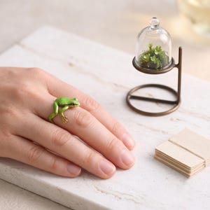 May include: A bright green frog ring featuring a realistic amphibian design that wraps around the finger. Beside it sits a small clear glass cloche containing tiny green plants, supported by a thin bronze-coloured metal stand on a base.