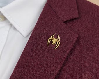 Pode incluir: Um alfinete de lapela em forma de aranha dourada preso à lapela de um blazer bordô texturizado. O blazer é usado sobre uma camisa branca de colarinho com um botão visível. O alfinete apresenta um design estilizado e simétrico.