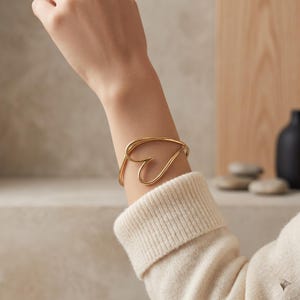 May include: A gold-toned metal cuff bracelet featuring a continuous, fluid wire design that forms an open, stylized heart shape. The bracelet is worn on the wrist, showcasing a smooth, polished finish and a minimalist, open-frame construction.