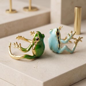 May include: Two open-style rings shaped like frogs. One ring is finished in glossy green enamel with gold-tone metal accents, while the other is finished in light blue enamel with gold-tone metal accents. Both rings feature detailed frog limbs and black eyes.