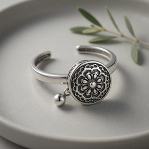 Könnte beinhalten: Verstellbarer Ring in Silber mit detailliertem Blumenmuster auf einer zentralen Scheibe. Unterhalb der Scheibe hängt eine kleine, baumelnde Kugel. Der Ring wird auf einer hellgrauen Oberfläche mit einem Zweig im Hintergrund präsentiert.