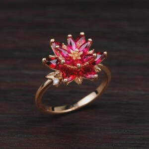 May include: A gold-colored ring featuring a flower design with red petals and a small diamond accent. The ring's petals are a vibrant red, and the band has a smooth, polished finish. The ring is set against a dark wooden background.