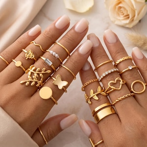 May include: A collection of gold-toned rings worn on fingers, featuring various designs including a rose, a butterfly, a heart, leaves, pearls, a rectangular stone, a plain circle, a twisted band, and a wide smooth band, creating a layered look.
