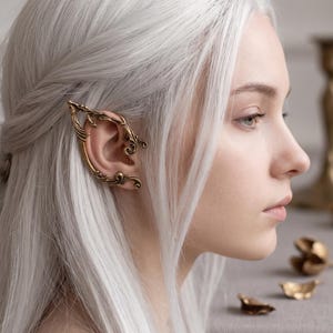 Può includere: Un ear cuff color oro decorato con intricati motivi a filigrana che avvolgono il bordo esterno dell'orecchio. L'accessorio in metallo presenta una finitura lucida con curve dettagliate e punte, progettato per agganciarsi saldamente alla cartilagine.