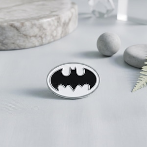 May include: An oval-shaped enamel pin featuring a black bat silhouette centred within a white background, enclosed by a thin silver-toned metal border. The design is clean and symmetrical, showcasing the iconic winged creature emblem in a classic high-contrast colour scheme.