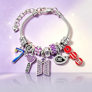 May include: A silver-toned charm bracelet featuring various pendants including a blue and yellow number 7, a purple heart with the text ARMY, a butterfly, a purple logo charm, a black whale-shaped charm with text, and three red circular charms.