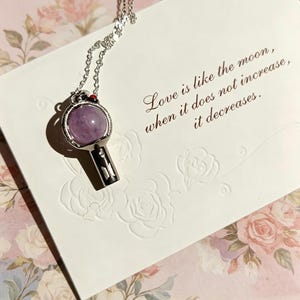 May include: A silver necklace with a purple gemstone pendant. The pendant has a black and silver handle design. The card reads, "Love is like the moon, when it does not increase, it decreases."