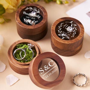 Könnte beinhalten: Vier Holz-Ringboxen mit transparenten Deckeln, jede mit einem Ring. Zwei Boxen haben den Text: "Paul & Emily July 7, 2025" und "S & C 12.10.2025". Eine Box hat den Buchstaben "H" und "03.01.25". Eine Box enthält zwei Ringe in grünem Moos. Ein silberner Ring ist ebenfalls sichtbar.