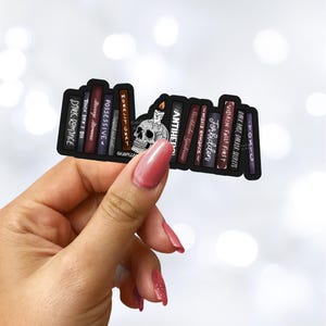 May include: A black sticker featuring a stack of books with titles like "Dark Romance" and "Toxic." A skull with a lit candle sits in front of the books. The sticker is held by a hand with pink nail polish.