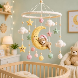 Op de afbeelding: Een gehaakte babymobiel met een grote gele maansikkel met een slapend gezicht, een kleine bruine teddybeer met een roze slaapmuts, witte wolken, pastelkleurige sterren en kleine bolletjes hangend aan een ring met houten kralen.