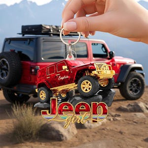 Op de afbeelding: Rode Jeep sleutelhanger met gouden accenten, met de naam "Sophia" en de tekst "Jeep girl". De sleutelhanger wordt voor een rode Jeep SUV gehouden. De achtergrond toont een bergketen.