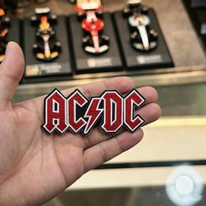 May include: An embroidered AC/DC patch featuring red lettering with a central red lightning bolt, outlined in black and white. The patch has a textured fabric surface and is shaped to follow the contours of the band's iconic logo design.