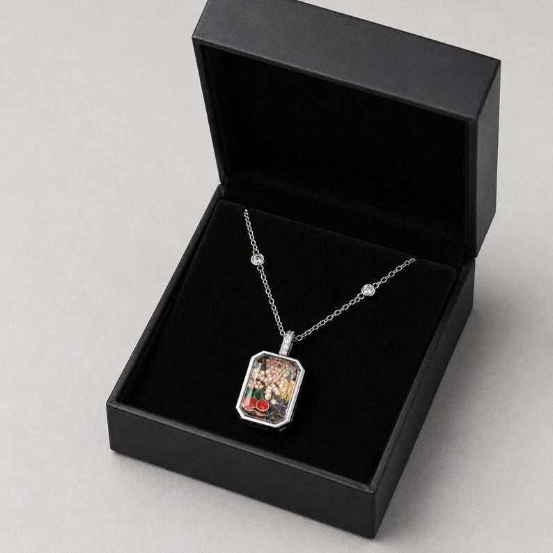 May include: A silver-toned rectangular pendant necklace featuring a clear glass front filled with small, colourful decorative charms including red cherries and yellow flowers, suspended on a delicate chain with two inset clear stones in a black box.