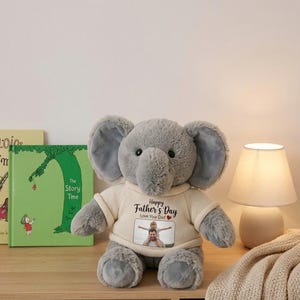 Puede incluir: Un elefante de peluche gris que lleva un suéter color crema con el texto "Happy Father's Day" y una foto. El elefante tiene orejas grandes y está sentado sobre una superficie de madera. Una lámpara y un libro están en el fondo.