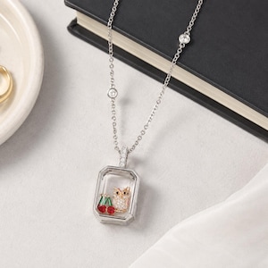 May include: A silver-toned necklace featuring a rectangular pendant with a clear glass front. Inside the pendant is a small, gold-toned owl figure with white stones and two red cherries with green stems. The chain includes small, round, clear stone accents.