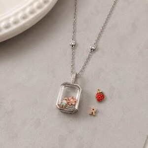 May include: A silver-toned locket necklace featuring a rectangular glass frame containing a small figure and a pink flower charm. The chain includes two small clear stones. Beside the locket are a red strawberry charm and a letter R charm.