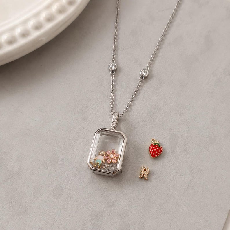May include: A silver-toned locket necklace featuring a rectangular glass frame containing a small figure and a pink flower charm. The chain includes two small clear stones. Beside the locket are a red strawberry charm and a letter R charm.