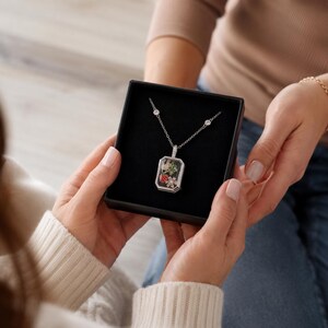 May include: A silver-toned necklace featuring a rectangular pendant with a clear front, containing small colourful charms of a strawberry, a green leaf, and a white flower. The chain includes two small round clear stones set along the silver links.